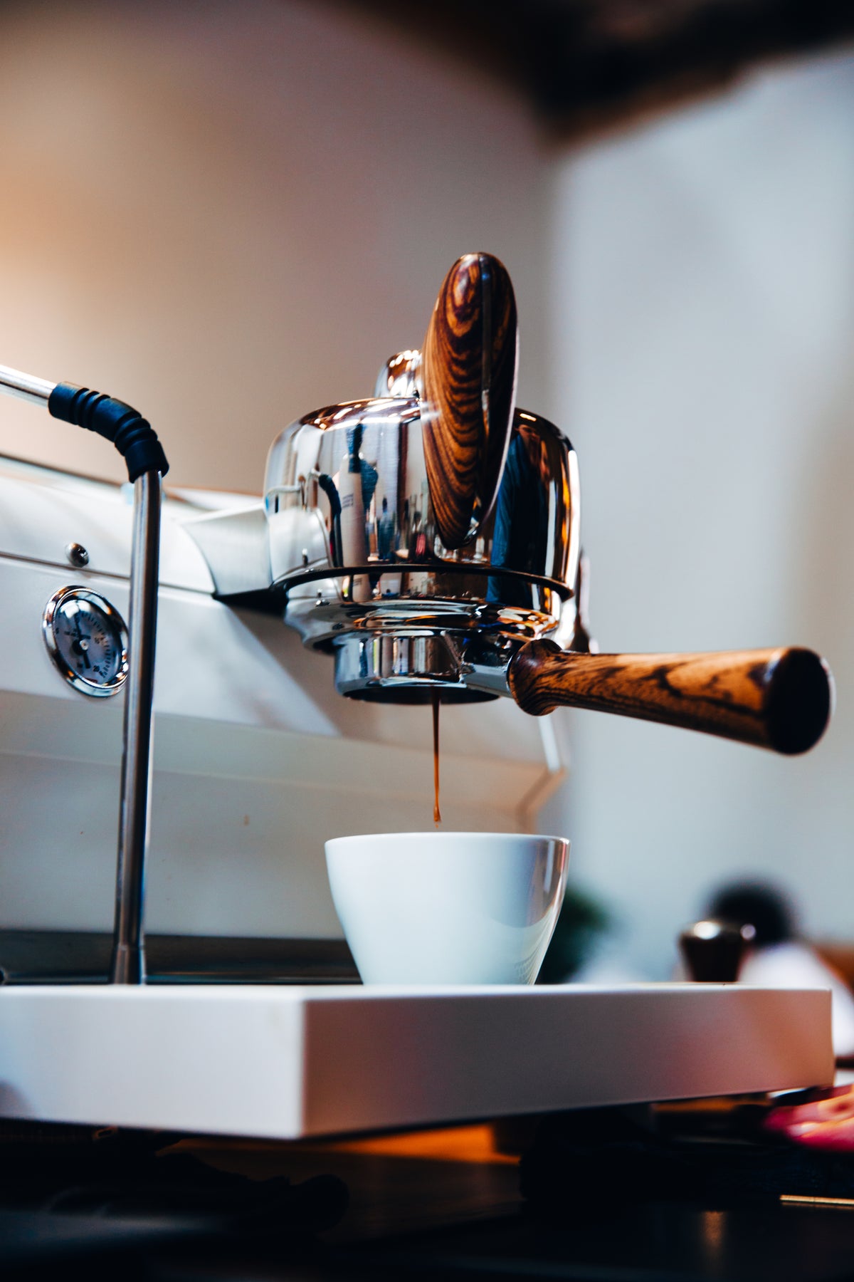 Close-up of an espresso machine with wood accents pouring espresso into a small white cup, with a warm blurred background